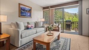 A living room with a sofa, coffee table, and a painting on the wall. There is a sliding glass door leading to an outdoor area with a barbecue grill. - Lichenhearth 09 (Snowmass Village)