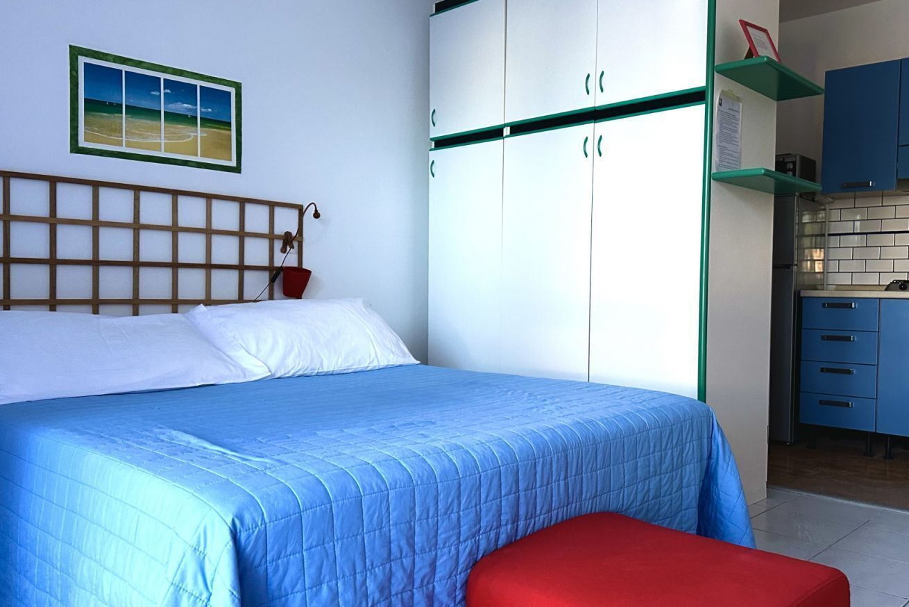 A bedroom with a bed, a red bench, a wardrobe, and a kitchen area.