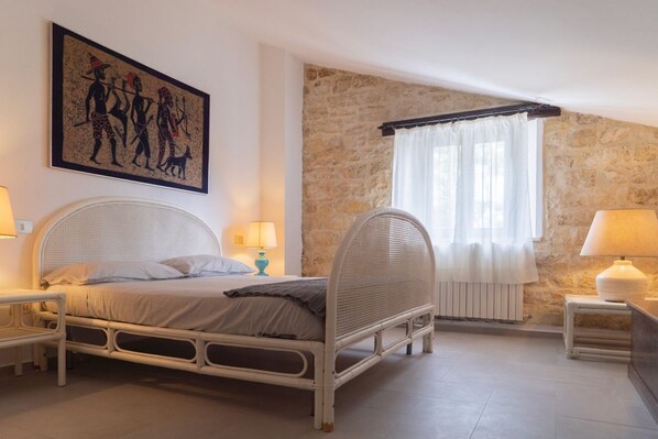 A bedroom with a stone wall, a bed with a woven headboard, a painting, and two bedside lamps.