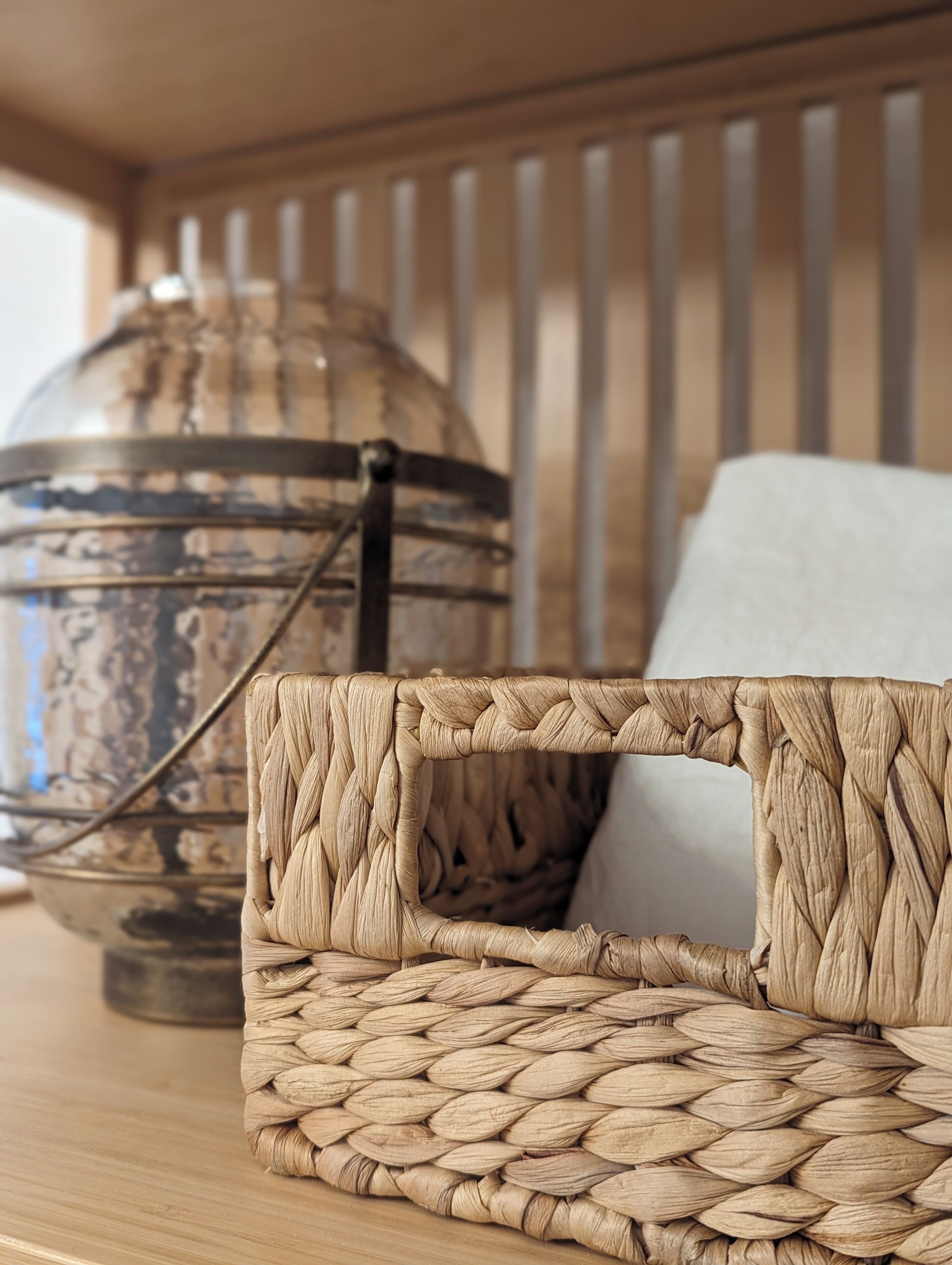 A woven basket with a handle, a decorative metal container, and a cushion on a wooden surface.