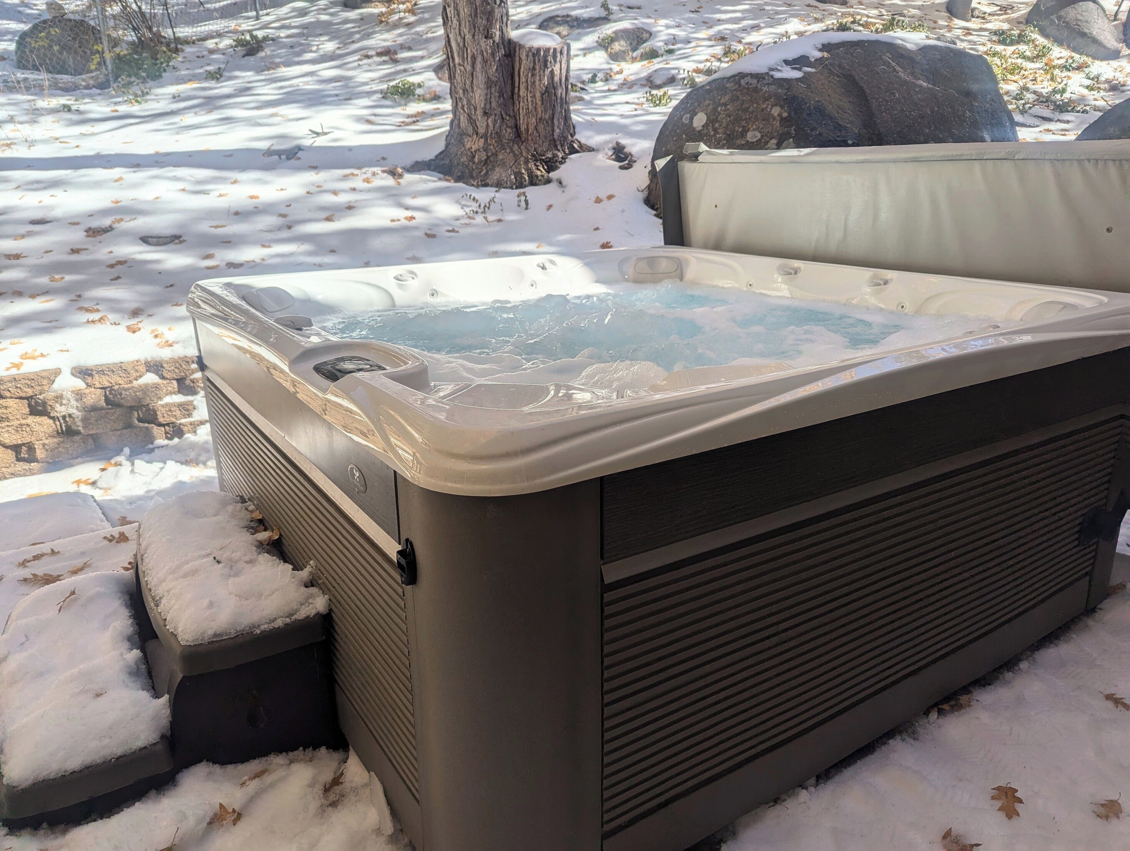 Ein Whirlpool vor verschneitem Gelände, einem Baumstumpf und einem Grill im Hintergrund.
