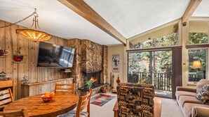 A cozy living room with a stone fireplace, wooden furniture, and a large window overlooking a balcony. - Lichenhearth 16 (Snowmass Village)