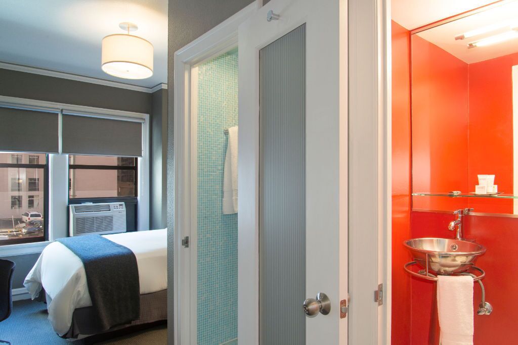 A hotel room with a bed, a window with an air conditioner, a bathroom with a red wall, a sink, and a towel rack.
