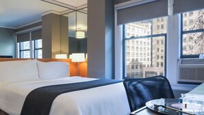 A hotel room with a bed, a desk with a laptop, a lamp, and a window with a city view. - Mini Full | City Access & Panoramic Views (Seattle)