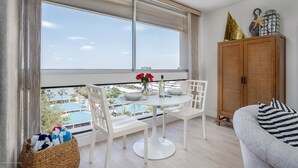 A modern living room with a round table and white chairs, a wicker basket, a wooden cabinet with decorative items, and a view of a pool and buildings through large windows. - Palm Bay Club 8th Floor On Complex Private Beach (Siesta Key)