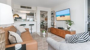 A modern living room with a sofa, armchair, and a TV displaying a beach scene. - Palm Bay Club 8th Floor On Complex Private Beach (Siesta Key)