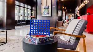 Un interior moderno con un panel azul perforado sobre una mesa redonda, una silla gris con cojín blanco y un mostrador rojo al fondo.