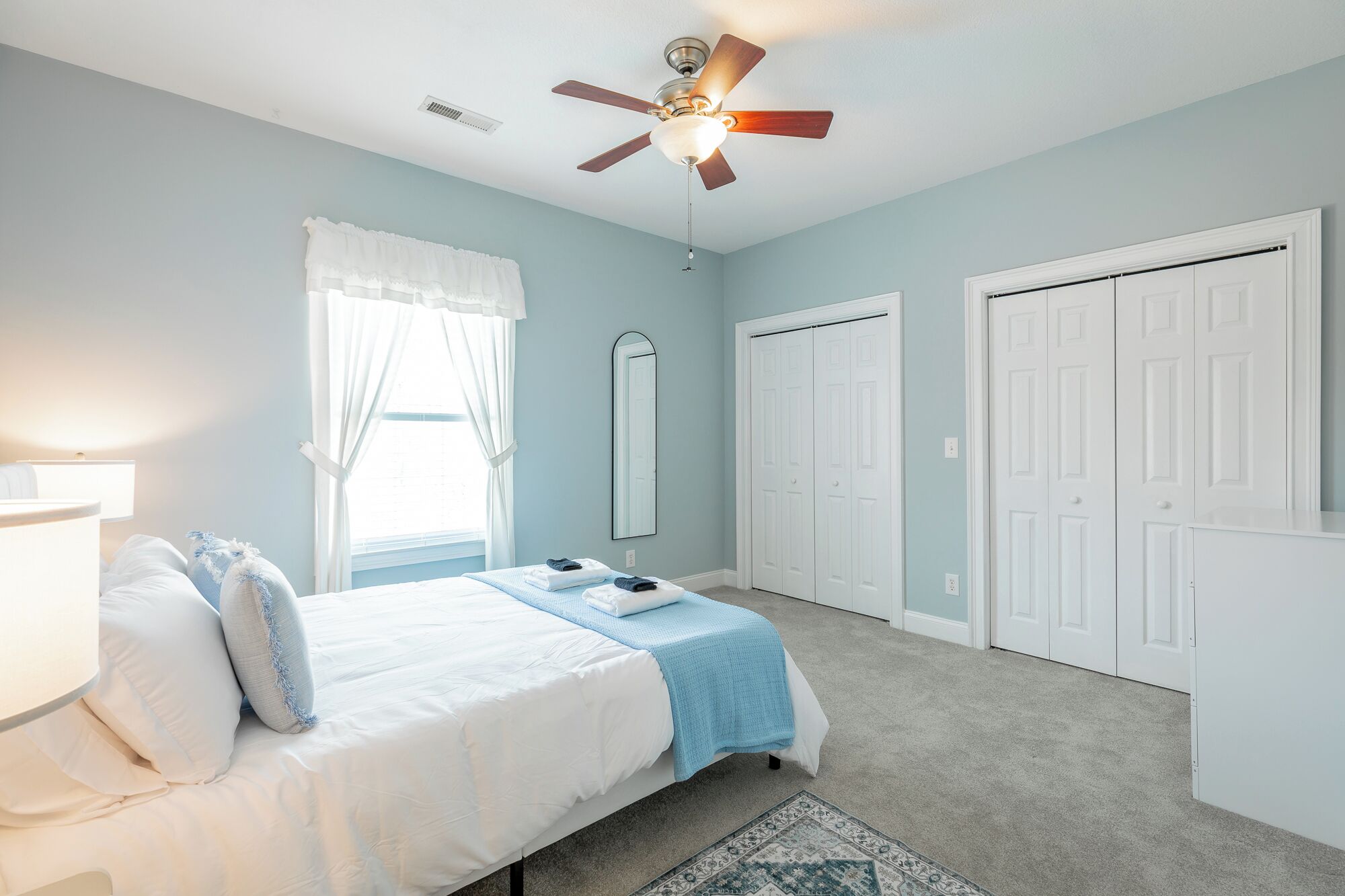Un dormitorio con cama, ventilador de techo, ventana con cortinas, espejo y armarios blancos.