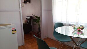 Un espace de vie compact comprenant un coin cuisine, un espace repas avec une table en verre et des chaises de couleur turquoise, une plante en pot et une fenêtre ornée de rideaux légers.