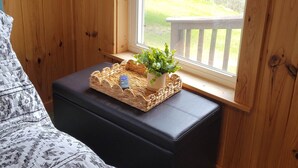 Een houten kamer met een bed, een kleintje tafeltje met een plant, en een raam met uitzicht op een houten terras en groen.