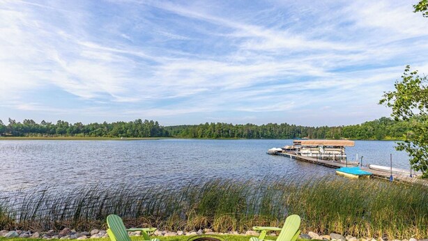A lakeside view with two green Adirondack chairs, a dock with boats, and a forested shoreline.