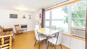 A room with a wooden table and chairs, a rocking chair, a map on the wall, and a view of a lake through large windows.