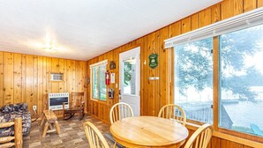 Ein Holzhäuschen im Inneren mit einem runden Holztisch und Stühlen, einer Couch, einem Kamin und einem großen Fenster mit Blick auf ein Gewässer.