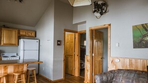 A compact kitchen with wooden cabinets, a white refrigerator, and a bar stool. There is a wooden door leading to another room.