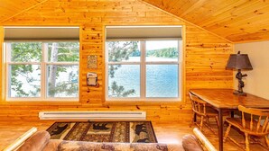 A cozy cabin with a large window overlooking a lake, wooden walls, a dining area, and a fireplace. - Cabin #6 - Loons' Nest (Lake Emma Township)