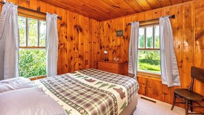 Un dormitorio de cabaña de madera con cama, cómoda y dos ventanas con cortinas.