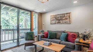 Uma sala de estar moderna com um sofá cinzento, almofadas coloridas, uma mesa de centro de madeira e uma grande janela com vista para o verde.