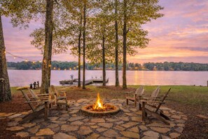 A stone fire pit surrounded by wooden chairs, a dock, and trees overlooking a lake at sunset. - Lake Oconee|Dock|Fire Pit|Big Views|Pet Friendly (Eatonton)
