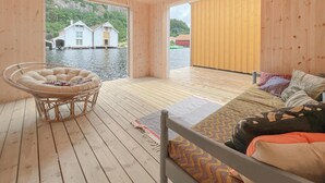 Un intérieur en bois avec un lit, une chaise en osier et une vue sur les maisons à travers une grande fenêtre.