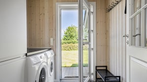 Uma lavanderia com máquina de lavar e secadora, um banco, e uma porta que dá para área externa com gramado e árvores.