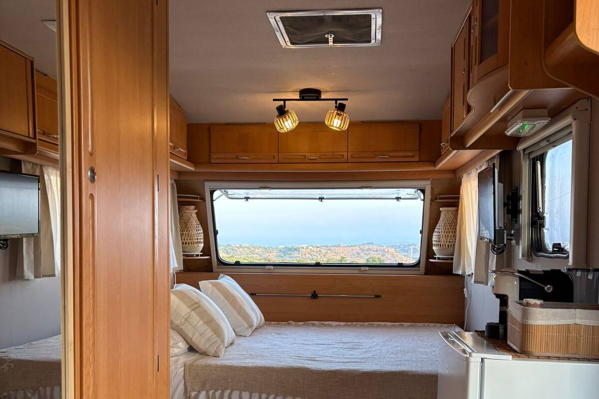 A compact RV interior with a bed, kitchenette, and a window providing a view of the landscape.