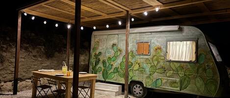 A covered outdoor area with a table and chairs, a camper van with cactus artwork, and string lights.