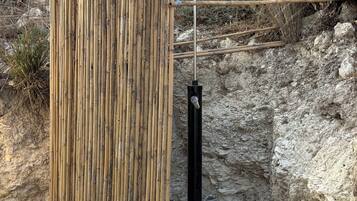 A bamboo fence with a metal structure behind it, set in a rocky outdoor area with gravel and sparse vegetation.