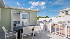 En terrasse med et bord og stole, en skydedør der fører til en balkon, og et hvidt hegn rundt om et poolområde.