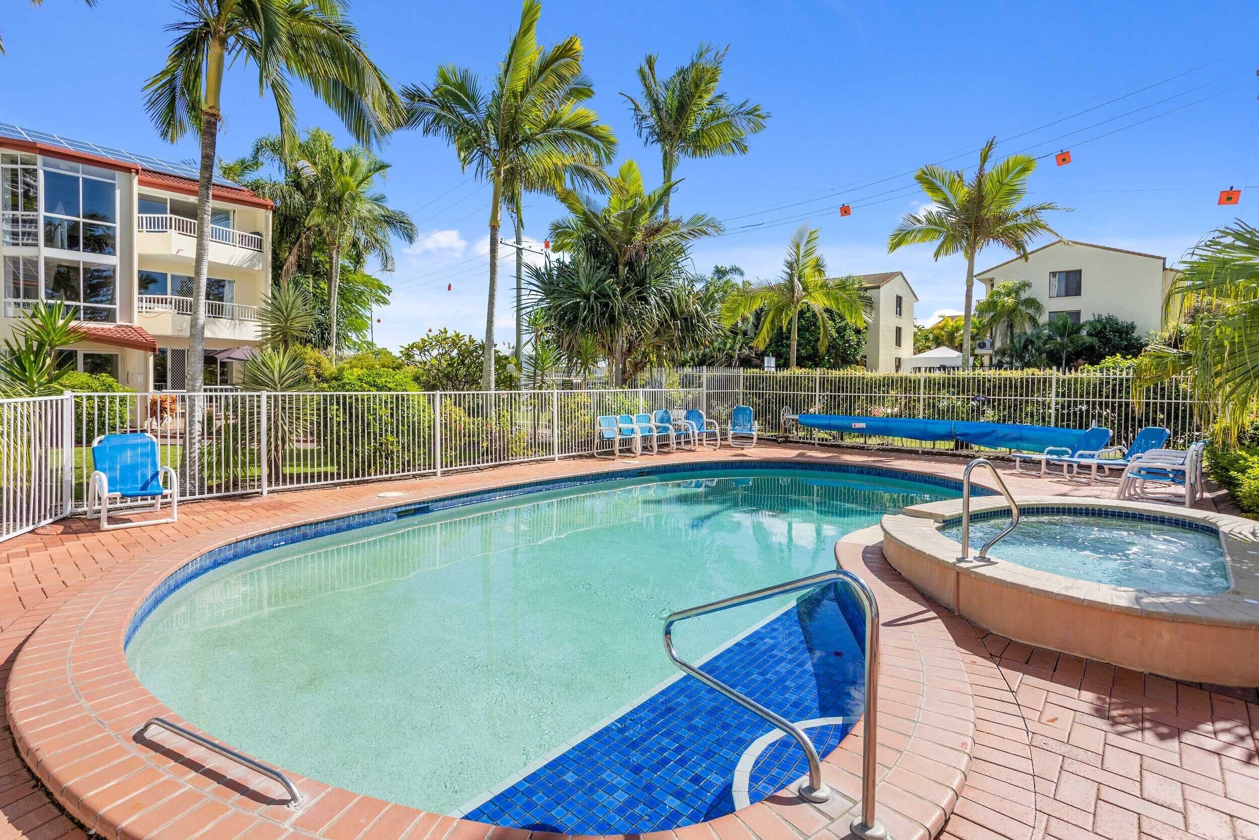 A swimming pool with a tiled edge, surrounded by a white fence, and palm trees in the background.