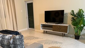 A living room with a grey sofa, a woven ottoman, a flat-screen TV on a white TV stand, a potted plant, and a sliding glass door.