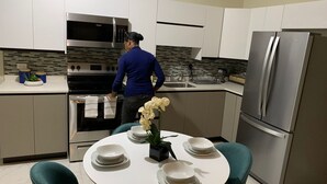 A modern kitchen with white cabinets, stainless steel appliances, and teal chairs around a white table with plates and a vase of flowers.