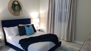 A bedroom with a large bed, a headboard, a nightstand, a lamp, a wall-mounted decoration, and a window with curtains.