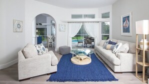 Una sala de estar moderna con dos sofás, una mesa de centro y un tapete azul. Hay una puerta de vidrio que da a un área exterior con piscina.