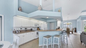 A modern kitchen with white cabinets, a central island with stools, and a dining area with a sofa and table. - Salty Sands - walk to Manasota Beach pool home (Englewood)