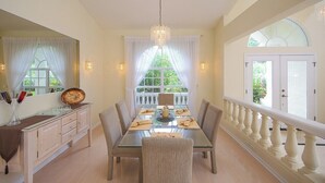 Un coin repas avec une table en verre, des chaises et un lustre. Une fenêtre avec des rideaux offre une vue sur la verdure à l’extérieur.