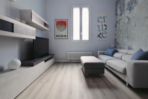 A modern living room with a flat-screen TV, a gray sofa, a white ottoman, and a radiator.
