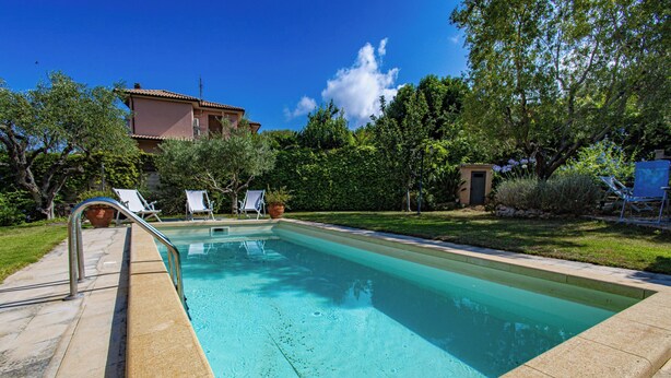 Una piscina rectangular con una terraza empavedada, rodeada de vegetación y un edificio al fondo.