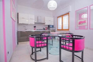 A modern kitchen with white cabinets, a glass dining table with pink chairs, and a large window with curtains.