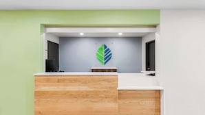 A reception desk with a wooden finish, a green wall to the left, and a grey wall with a leaf-shaped decoration above it. - Woodspring Suites Kansas City Airport (Kansas City)