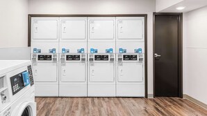A laundry facility with multiple washing machines and dryers, a wooden floor, and a dark door. - Woodspring Suites Kansas City Airport (Kansas City)