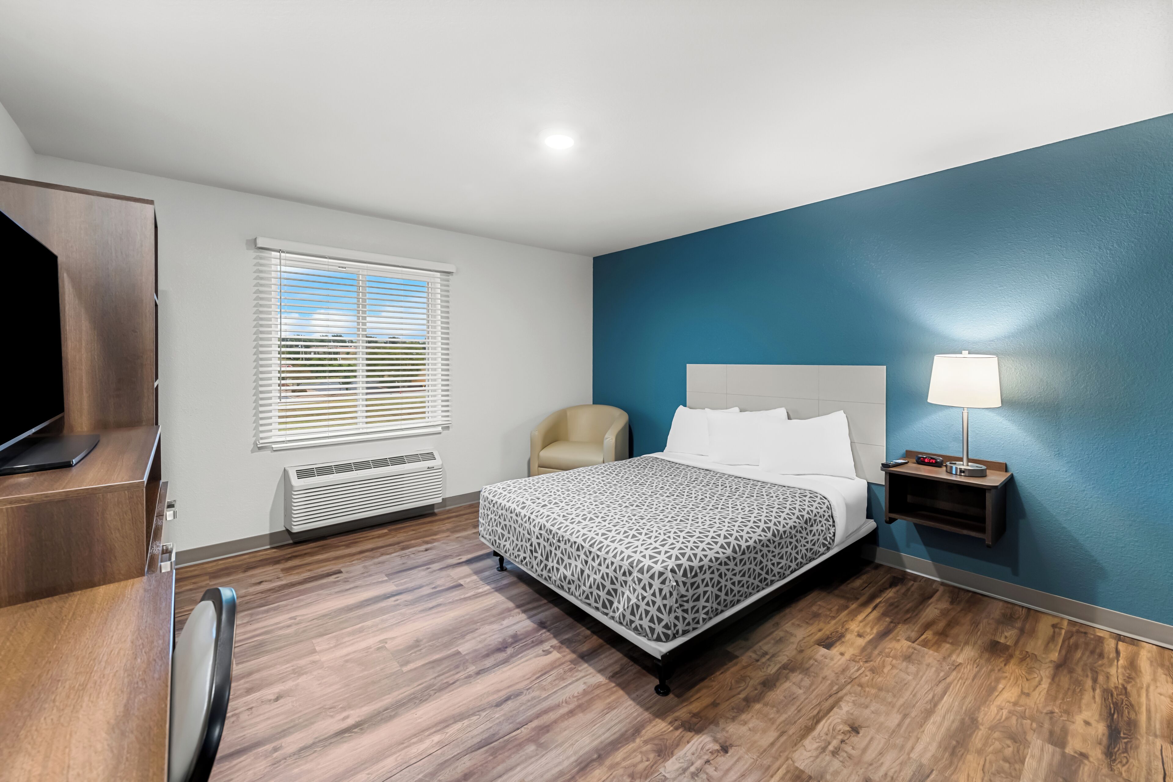 A modern hotel room with a wooden floor, a bed with patterned bedding, a chair, a desk with a flat-screen TV, a window with blinds, and a nightstand with a lamp.
