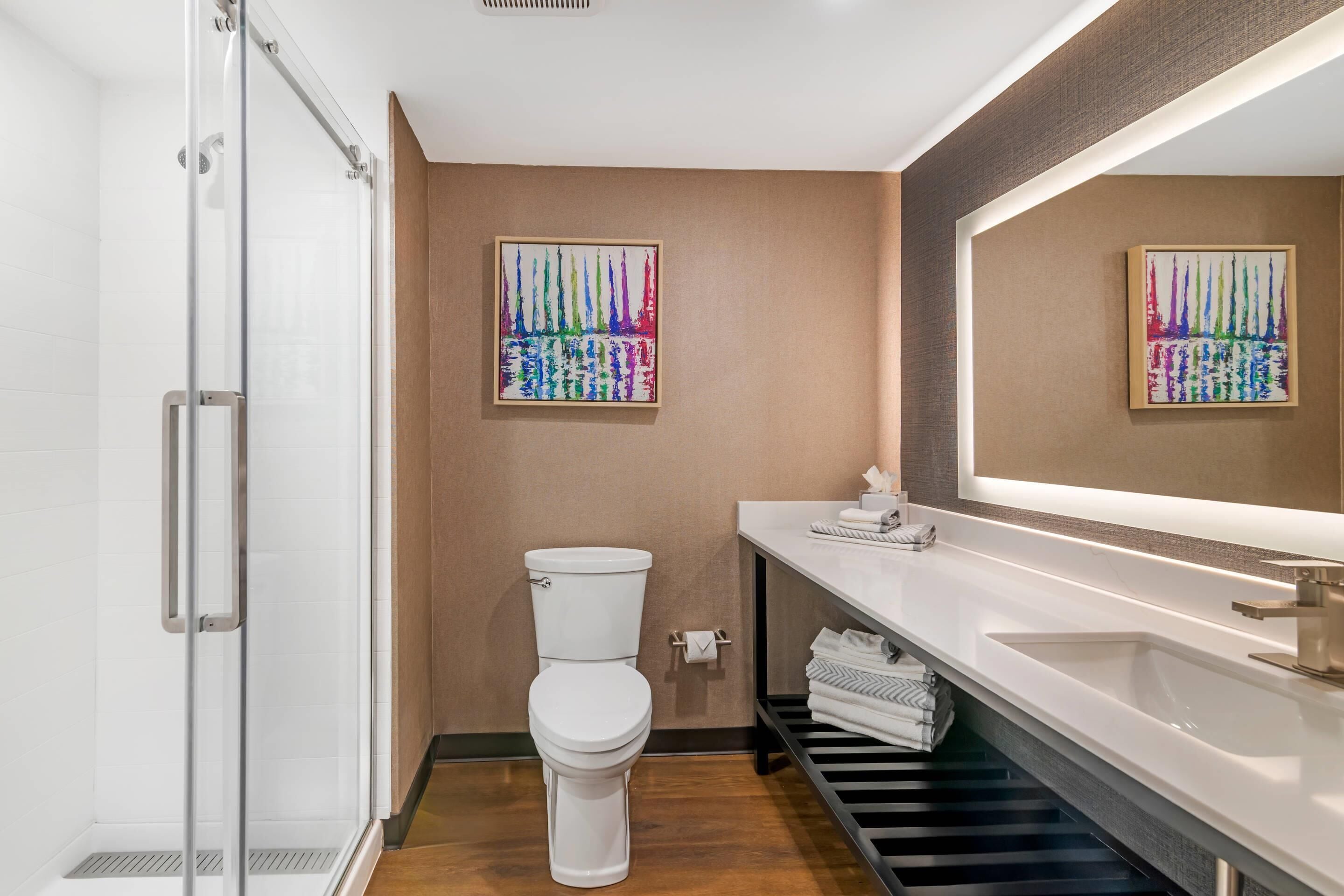 Uma casa de banho moderna com cabine de chuveiro em vidro, sanita branca, espelho grande e lavatório branco com um porta-toalhas.