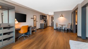 A modern hotel room with a wooden floor, a desk with an orange chair, a flat-screen TV, and a dining area with two chairs. - Cambria Hotel & Suites Plymouth (Plymouth)