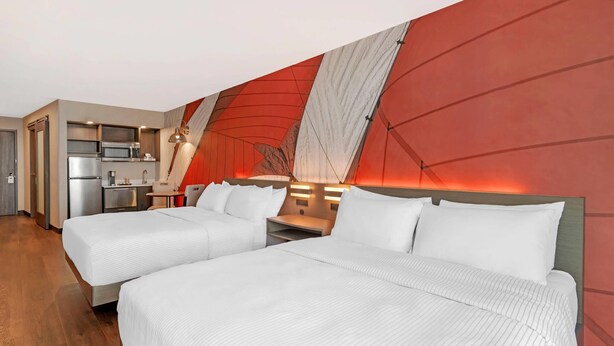 A hotel room with two beds, a kitchenette, and a red accent wall. - Cambria Hotel & Suites Plymouth (Plymouth)