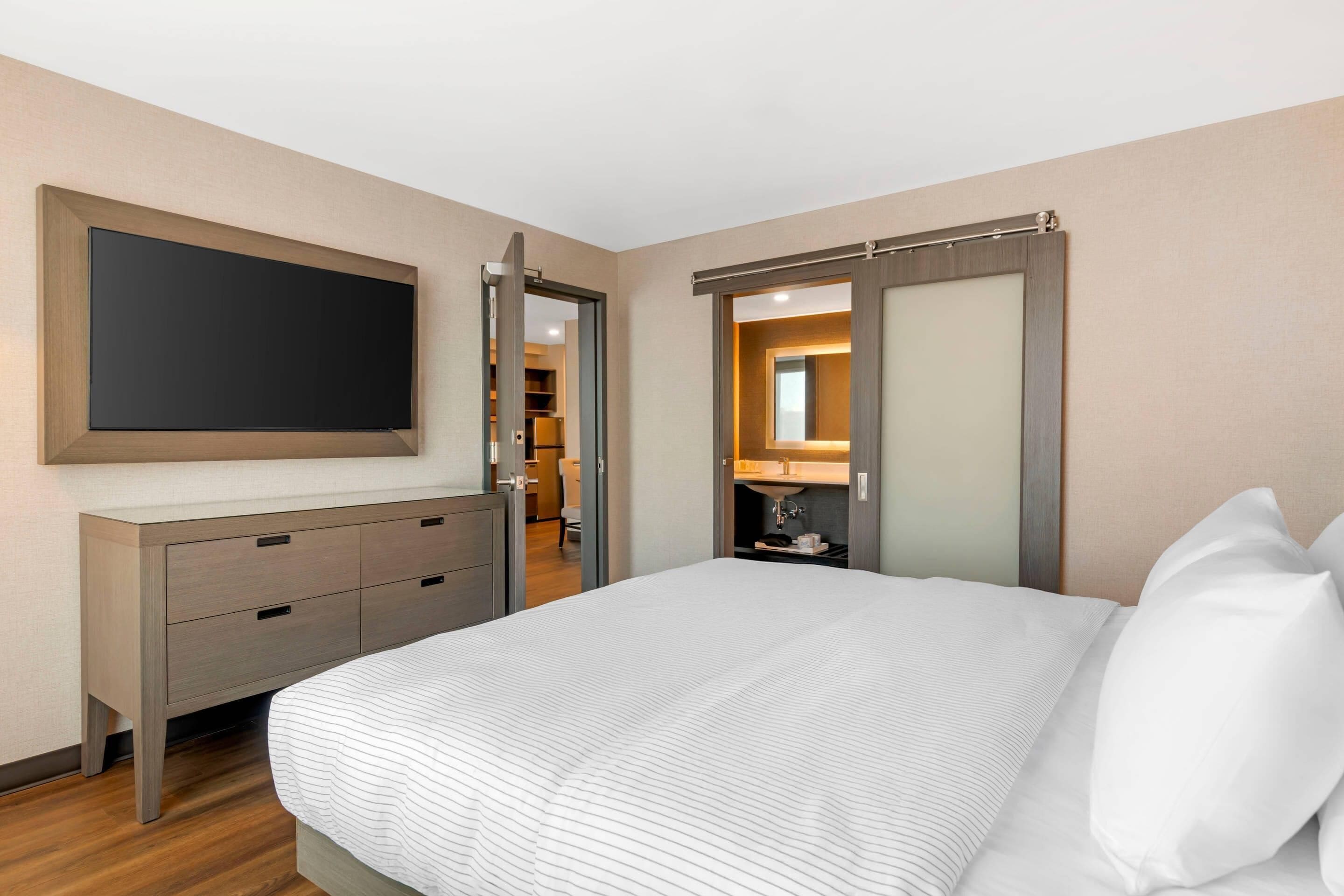 A hotel room with a bed, a TV, a dresser, and a bathroom visible through an open door.