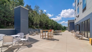 An outdoor patio area with tables and chairs, a stone wall, and a building with large windows. - Cambria Hotel & Suites Plymouth (Plymouth)