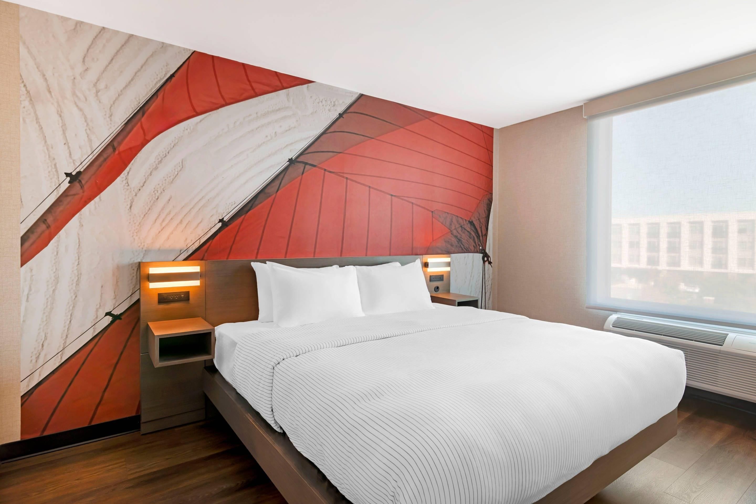 Una habitación de hotel moderna con una cama grande, una mesita de noche con una lámpara, un calentador de pared y un ventanal con vistas a la ciudad.