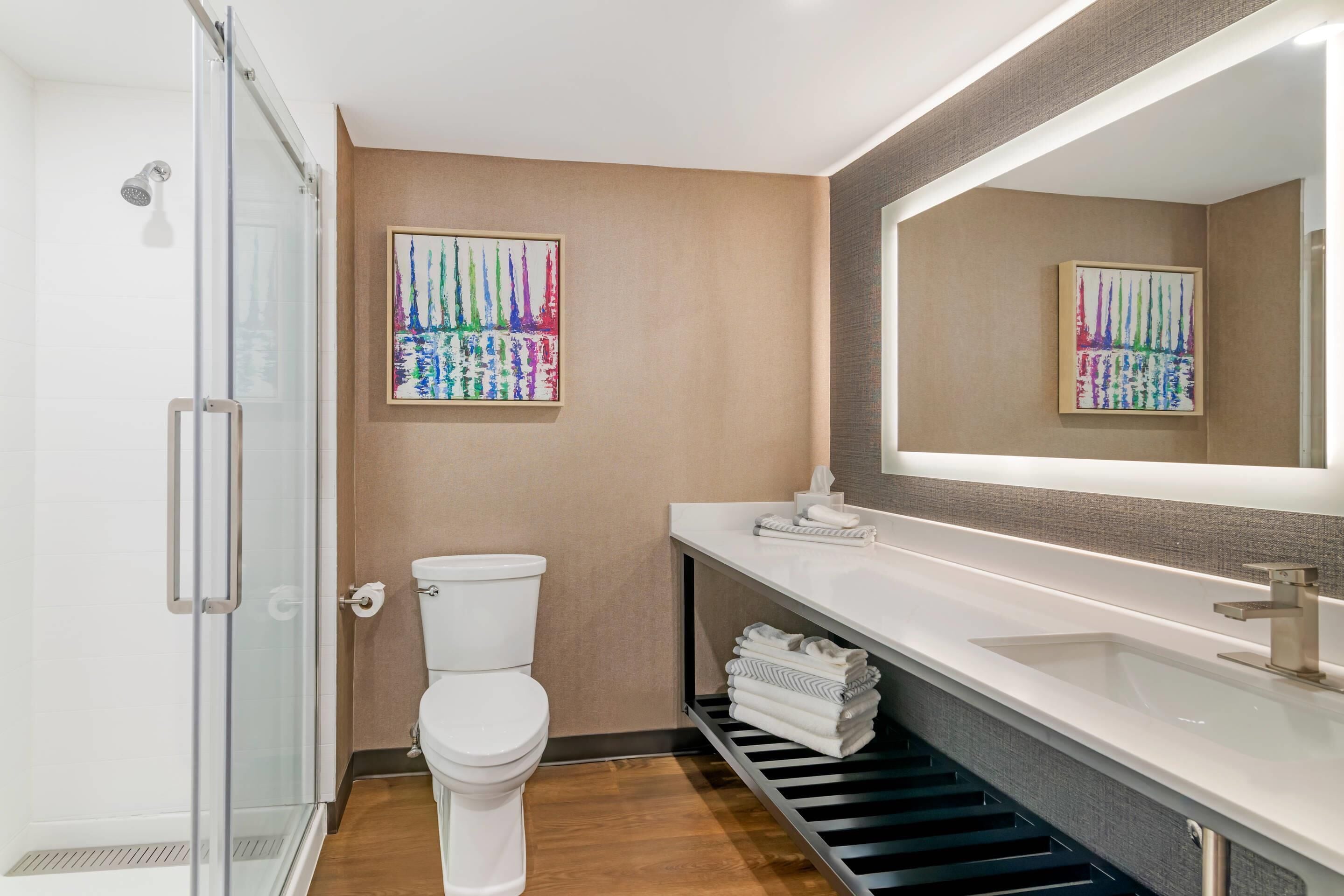 Un baño moderno con cabina de ducha de vidrio, inodoro, lavamanos con espejo y dos cuadros abstractos coloridos en la pared.