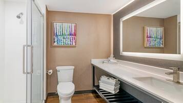 Un baño moderno con cabina de ducha de vidrio, inodoro, lavamanos con espejo y dos cuadros abstractos coloridos en la pared.