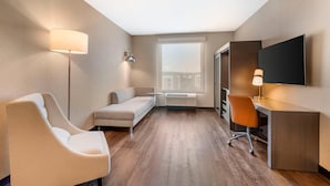 A modern hotel room with a sofa, a desk, a chair, a TV, and a large window. - Cambria Hotel & Suites Plymouth (Plymouth)